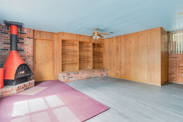 unfurnished living room with wooden walls, ceiling fan, a wood stove, baseboards, and wood finished floors