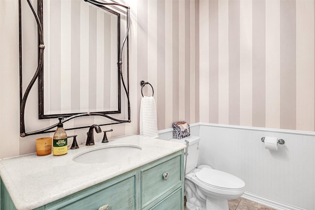 half bath featuring toilet, wallpapered walls, wainscoting, and vanity