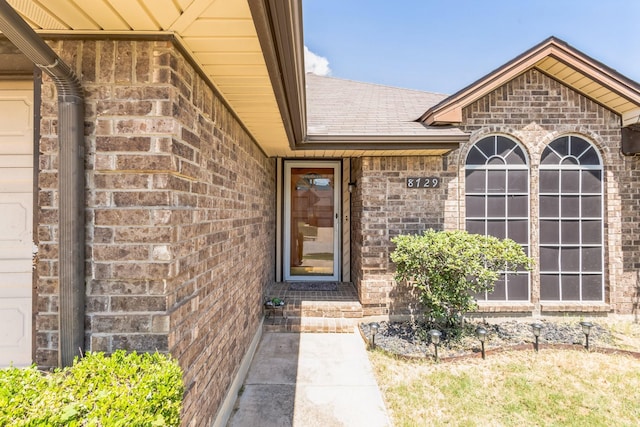 view of exterior entry with roof with shingles and brick siding