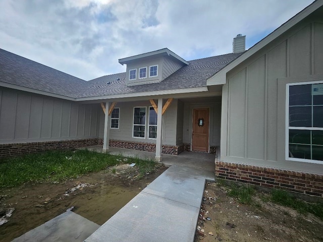 entrance to property with covered porch, a chimney, brick siding, a shingled roof, and board and batten siding