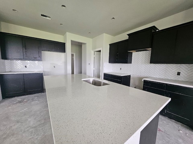kitchen featuring dark cabinetry, an island with sink, backsplash, and light stone countertops