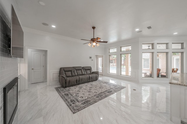 living room with a decorative wall, crown molding, a fireplace, a ceiling fan, and a wainscoted wall