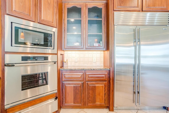 kitchen featuring backsplash, a warming drawer, light stone countertops, glass insert cabinets, and built in appliances