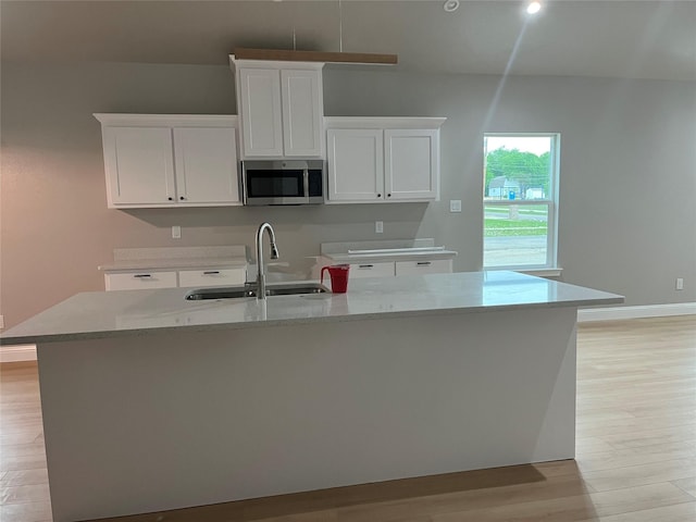 kitchen with a kitchen island with sink, stainless steel microwave, white cabinets, a sink, and light wood-style flooring