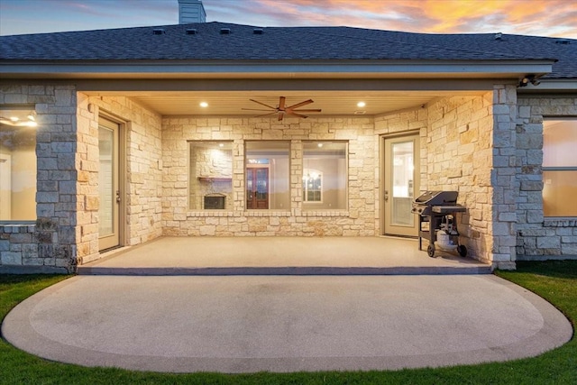 entrance to property featuring a patio, a ceiling fan, roof with shingles, and stone siding