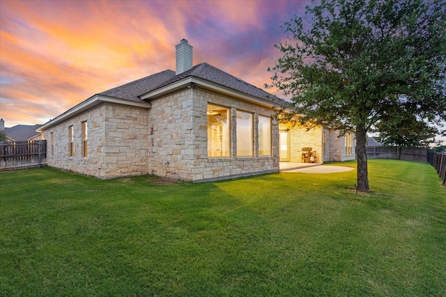 back of property at dusk featuring a chimney, a fenced backyard, a lawn, and stone siding