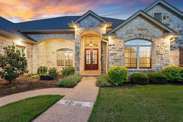 exterior entry at dusk with a lawn, roof with shingles, and stone siding