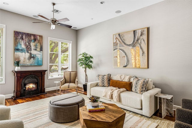 living room featuring ceiling fan, visible vents, wood finished floors, baseboards, and a warm lit fireplace