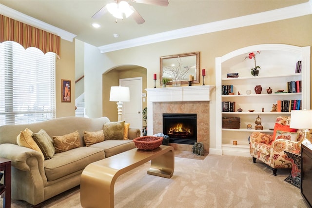 carpeted living area with ornamental molding, a ceiling fan, a tile fireplace, stairway, and arched walkways