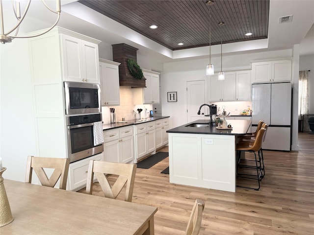 kitchen featuring stainless steel dishwasher, dark countertops, a tray ceiling, a sink, and light wood finished floors