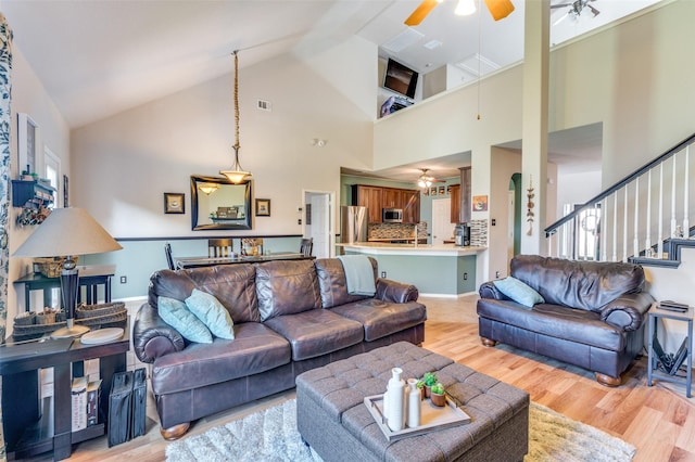 living area featuring ceiling fan, high vaulted ceiling, light wood-style flooring, visible vents, and stairway