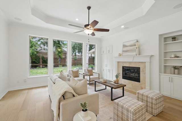living room with light wood-type flooring, a raised ceiling, a premium fireplace, ceiling fan, and built in shelves