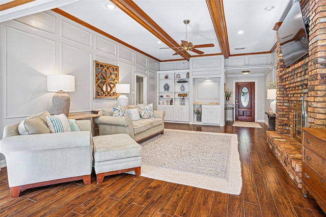 living area featuring ornamental molding, ceiling fan, dark wood-style flooring, a brick fireplace, and beam ceiling