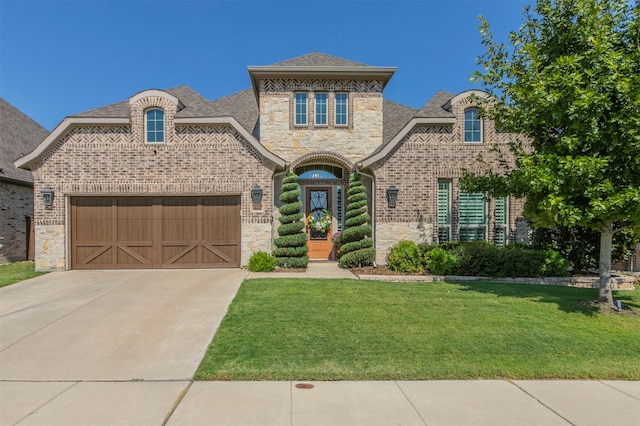 french provincial home with brick siding, a front yard, an attached garage, concrete driveway, and stone siding