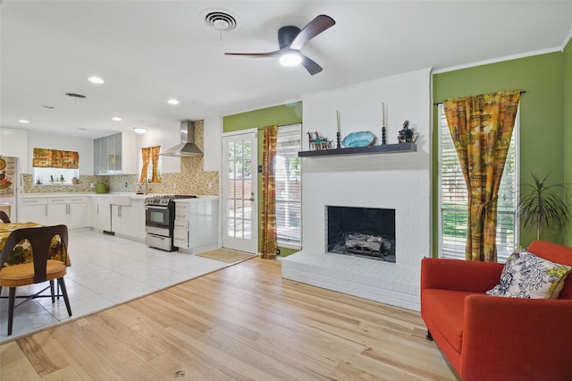 living area with a brick fireplace, light wood-type flooring, visible vents, ceiling fan, and ornamental molding