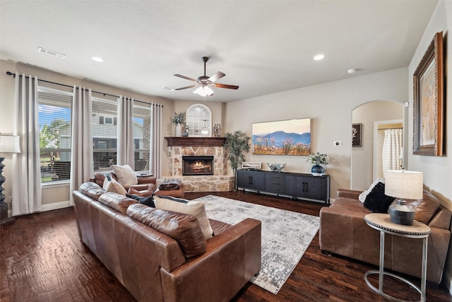 living room featuring a ceiling fan, dark wood finished floors, visible vents, a stone fireplace, and arched walkways