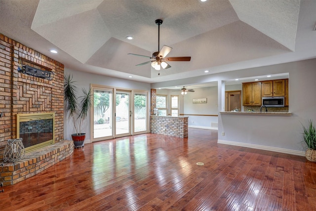 unfurnished living room featuring a raised ceiling, a ceiling fan, a brick fireplace, baseboards, and wood finished floors