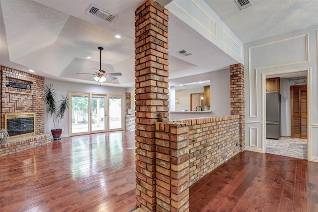 unfurnished living room featuring a textured ceiling, hardwood / wood-style flooring, a tray ceiling, a brick fireplace, and ceiling fan