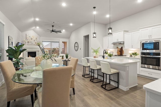 dining area with light wood-style floors, vaulted ceiling, a warm lit fireplace, and a ceiling fan