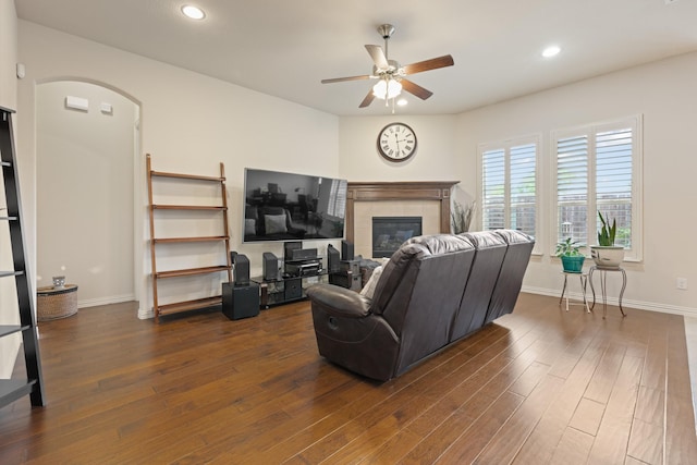 living area featuring baseboards, a ceiling fan, a tiled fireplace, and dark wood-type flooring