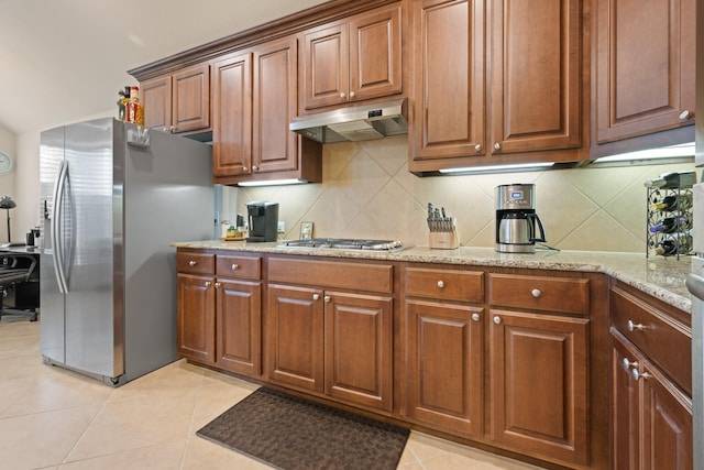 kitchen with stainless steel appliances, brown cabinets, light stone counters, under cabinet range hood, and backsplash