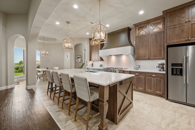 kitchen featuring stainless steel fridge with ice dispenser, a chandelier, premium range hood, tasteful backsplash, and a kitchen island with sink