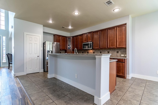 kitchen featuring stainless steel appliances, tasteful backsplash, light stone counters, visible vents, and an island with sink