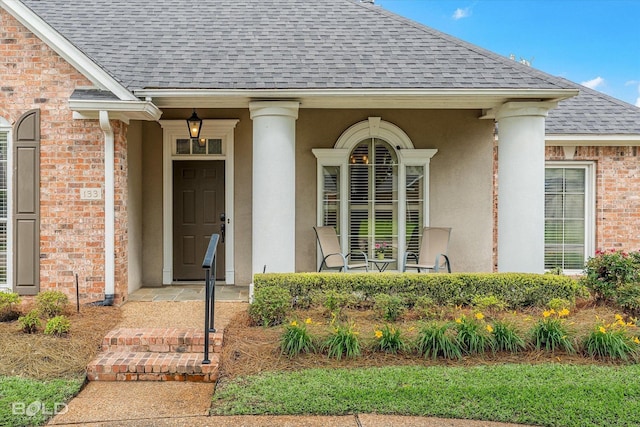 view of exterior entry with covered porch, stucco siding, roof with shingles, and brick siding