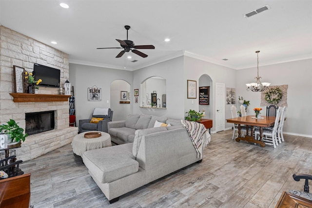 living room featuring crown molding, ceiling fan with notable chandelier, a stone fireplace, and visible vents