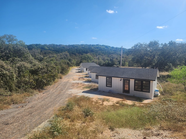view of front of property featuring a wooded view, roof with shingles, and dirt driveway