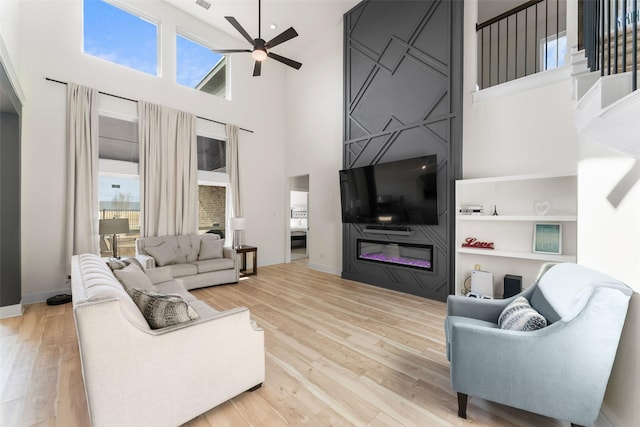 living room with a large fireplace, a ceiling fan, wood finished floors, and a towering ceiling