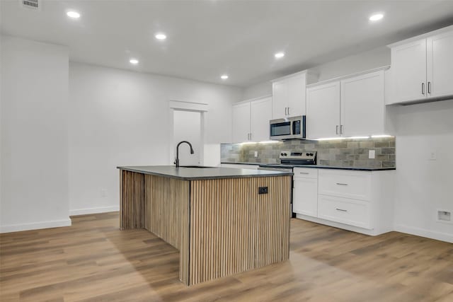 kitchen featuring stainless steel appliances, a kitchen island with sink, dark countertops, and backsplash
