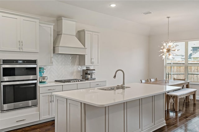 kitchen with stainless steel appliances, custom exhaust hood, a sink, a center island with sink, and light countertops