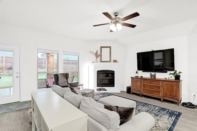 living room with a ceiling fan, light wood-type flooring, baseboards, a glass covered fireplace, and vaulted ceiling