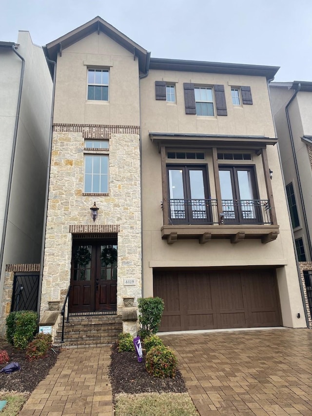 view of front of home with an attached garage, french doors, stone siding, a balcony, and stucco siding