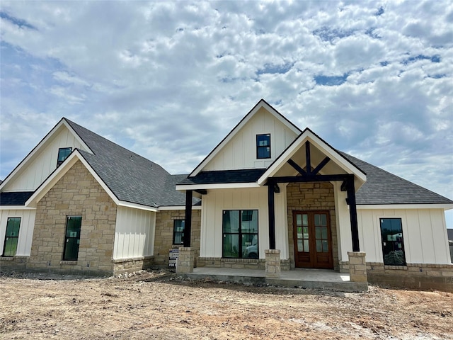 view of front of property featuring board and batten siding, french doors, covered porch, a shingled roof, and stone siding