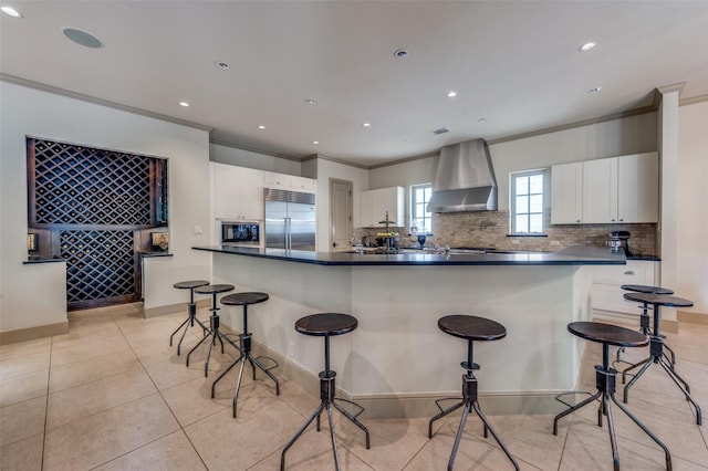 kitchen featuring crown molding, wall chimney exhaust hood, built in appliances, and a breakfast bar