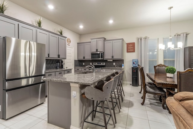 kitchen with an inviting chandelier, light stone countertops, gray cabinetry, appliances with stainless steel finishes, and decorative backsplash