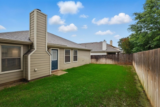 rear view of house with a shingled roof, a chimney, a lawn, and a fenced backyard