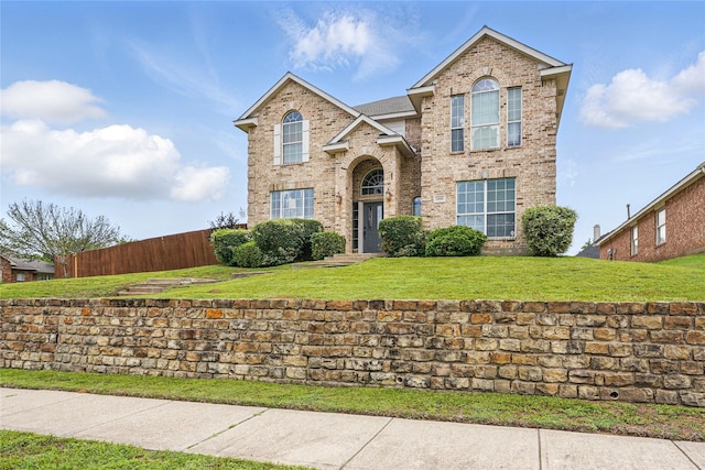 traditional-style home featuring a front lawn, a shingled roof, brick siding, and fence