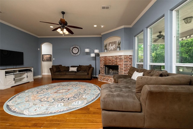 living area featuring wood finished floors, a brick fireplace, crown molding, visible vents, and ceiling fan