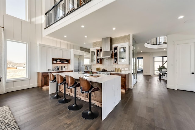1725 Ranch House Rd, Celina, TX 75009 - kitchen featuring wall chimney range hood, dark wood finished floors, light countertops, an island with sink, and glass insert cabinets