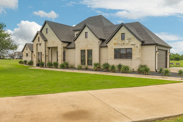 french country style house featuring an attached garage, stone siding, a front lawn, brick siding, and roof with shingles