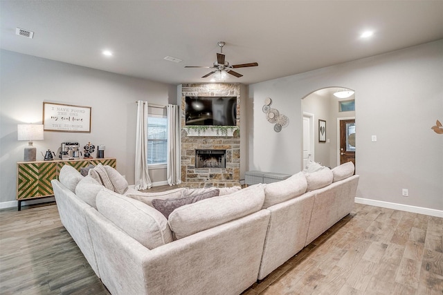 living room with visible vents, ceiling fan, light wood finished floors, arched walkways, and a stone fireplace