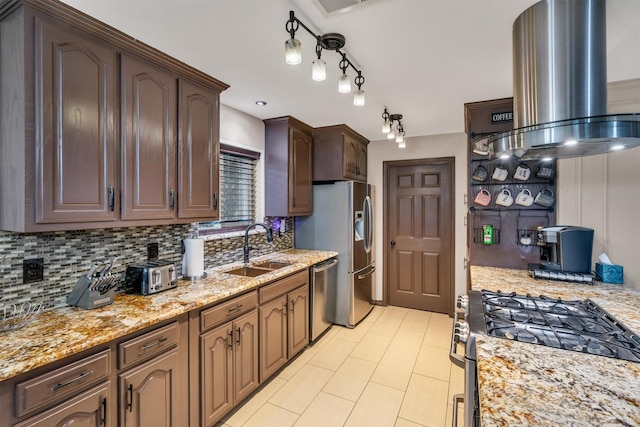 kitchen with appliances with stainless steel finishes, a sink, island range hood, decorative backsplash, and light stone countertops