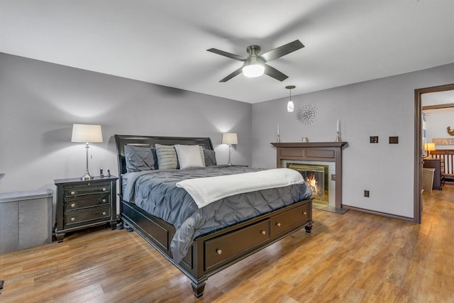bedroom with wood finished floors, a glass covered fireplace, baseboards, and a ceiling fan