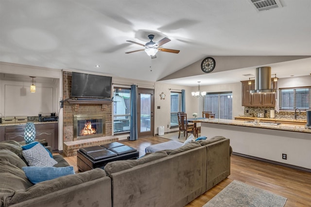 living room featuring a ceiling fan, a fireplace, light wood finished floors, vaulted ceiling, and a chandelier