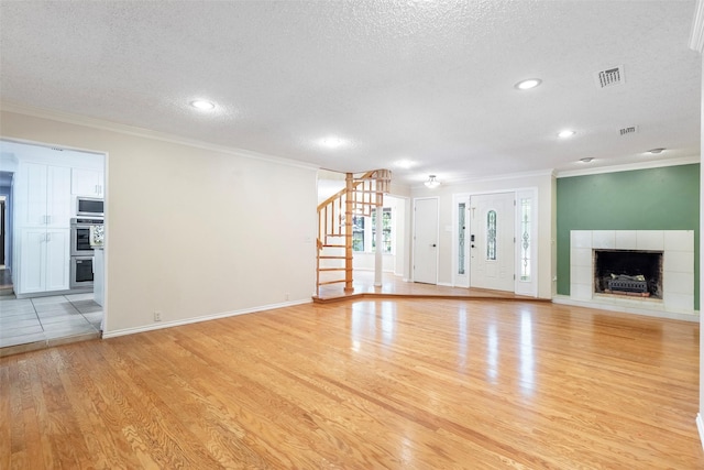 unfurnished living room with a textured ceiling, light wood-style flooring, ornamental molding, a tile fireplace, and stairway