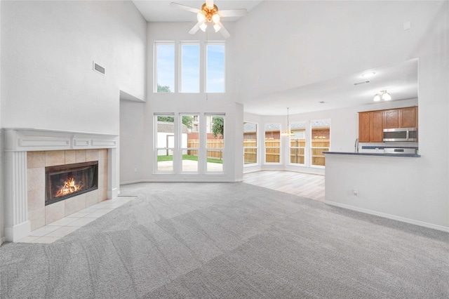 unfurnished living room featuring a ceiling fan, light carpet, a tiled fireplace, a towering ceiling, and baseboards