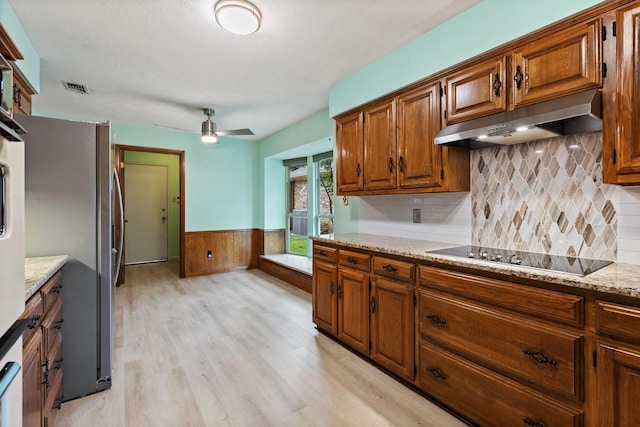 kitchen with under cabinet range hood, freestanding refrigerator, black electric stovetop, light wood-style flooring, and brown cabinetry